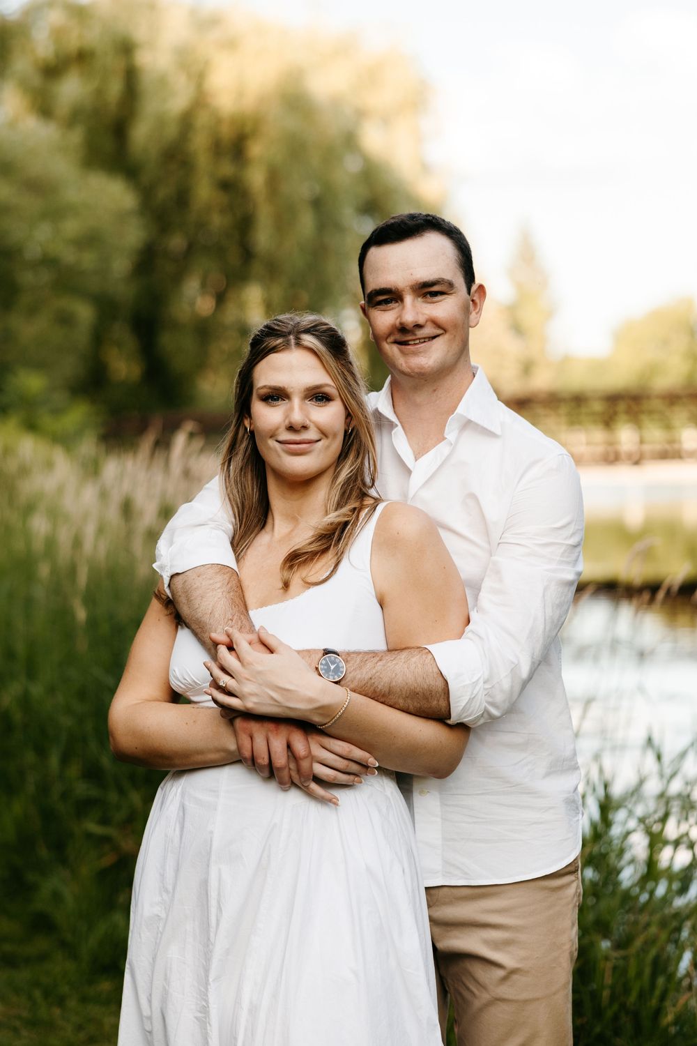 Couple embracing outdoors near a river, woman in white dress, man in white shirt and khaki pants, surrounded by lush greenery.