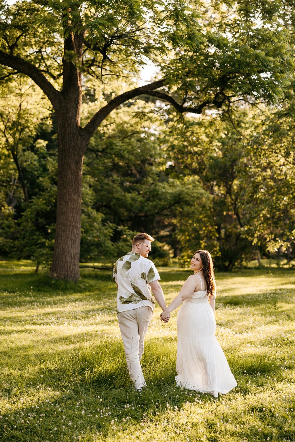 A couple holds hands walking through a sunlit green park, the woman in a white dress and the man in a floral shirt.