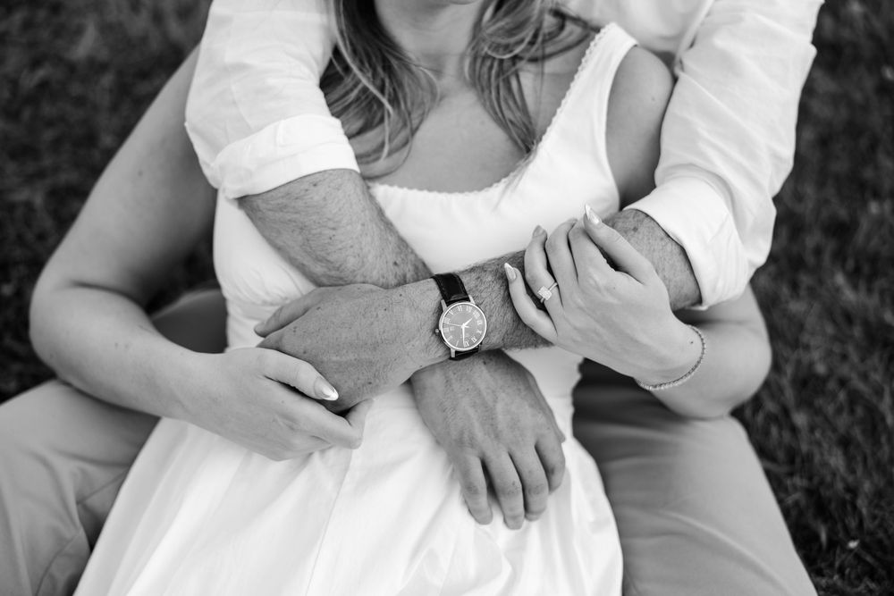 Black and white photo of a couple embracing, man wrapping arms around woman in white dress outdoors.