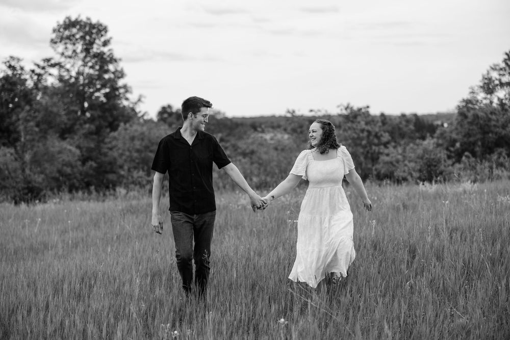 Black and white photo of a couple holding hands and walking through a grassy field with trees in the background.