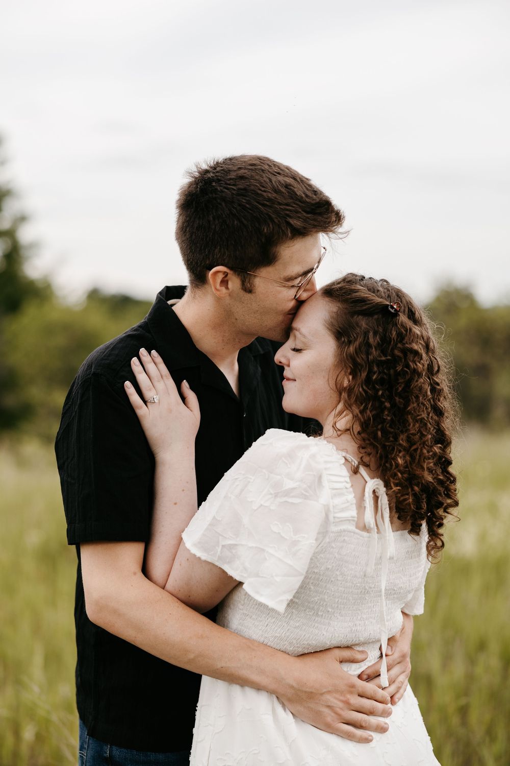 A couple shares a tender moment in a green field, man in black shirt kisses woman in white dress on the forehead.