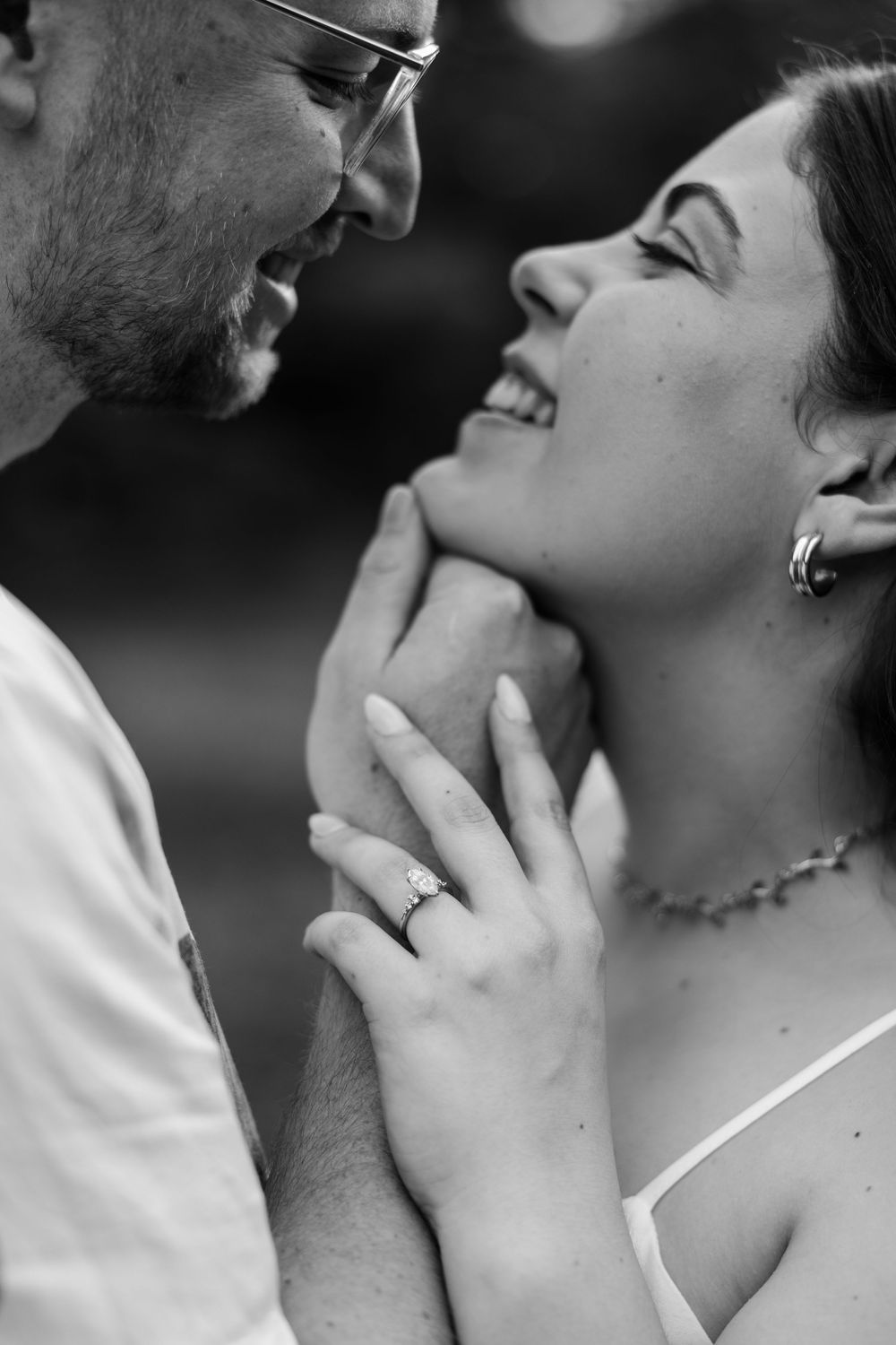 Black and white photo of a smiling couple almost kissing, woman wearing engagement ring and delicate necklace.