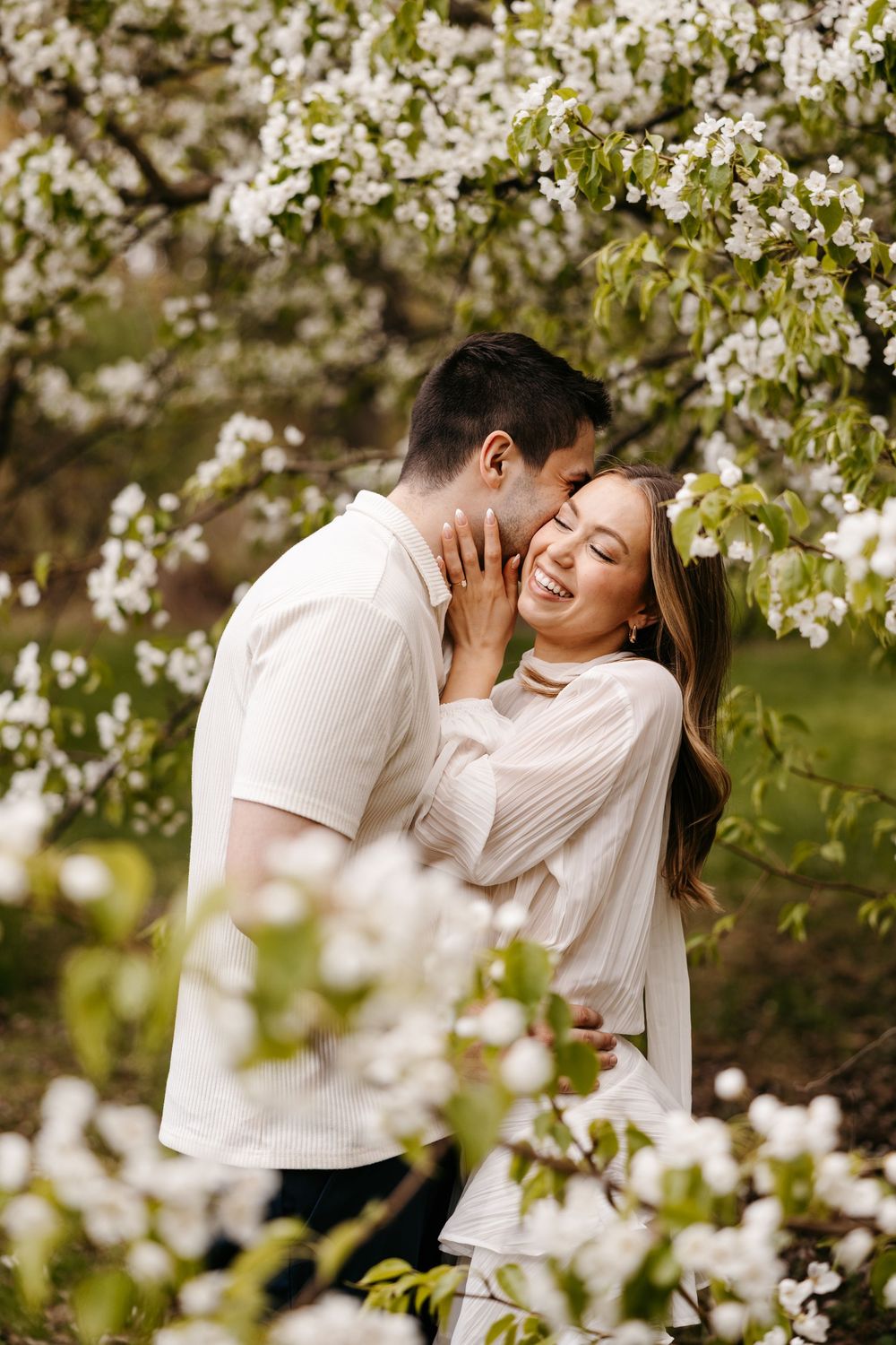 Couple embracing and laughing among white blooming cherry trees in spring, wearing matching white outfits.