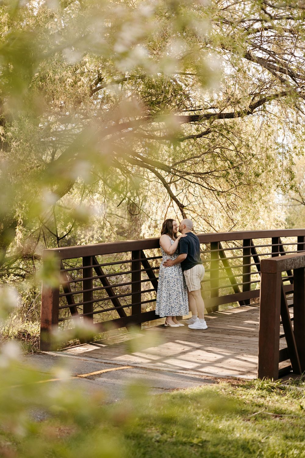 Couple embracing on a wooden bridge surrounded by lush green weeping willow trees in soft natural light.