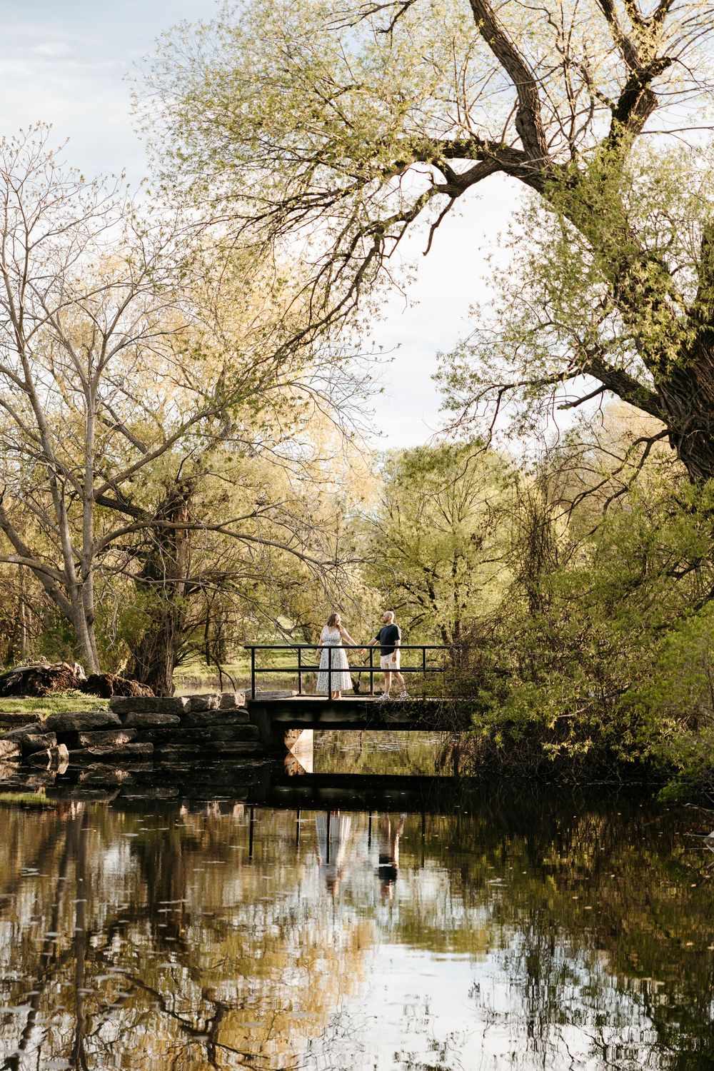 Couple standing on a rustic stone bridge surrounded by lush trees with reflections in calm water below.