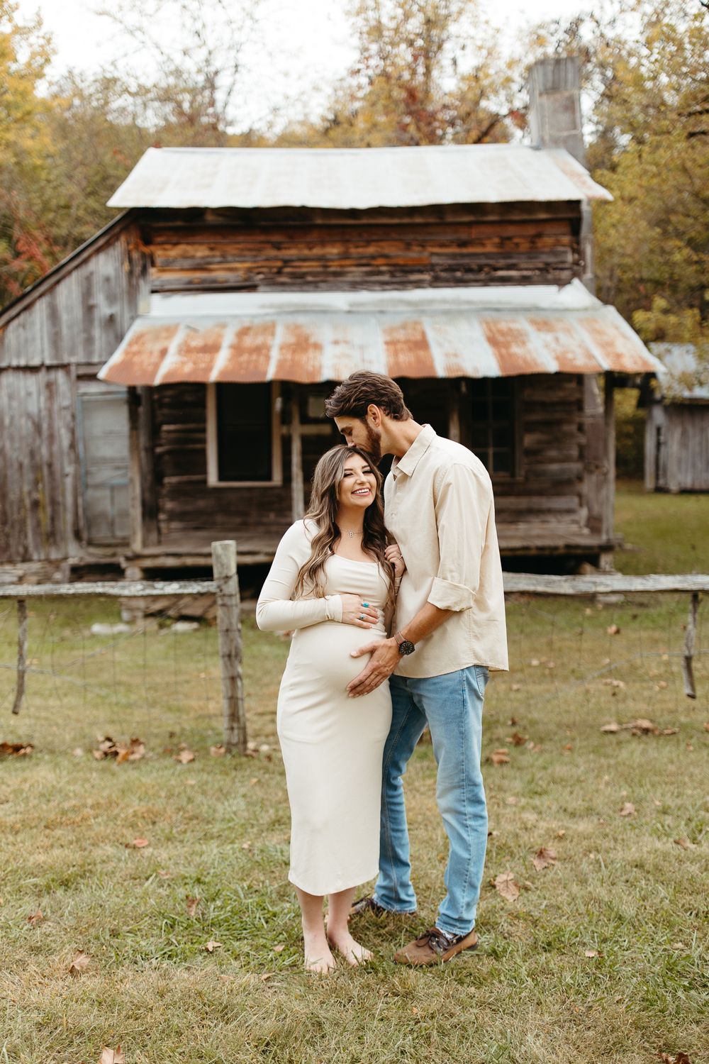 Expecting couple poses in front of rustic cabin wearing casual clothing during autumn photo session.