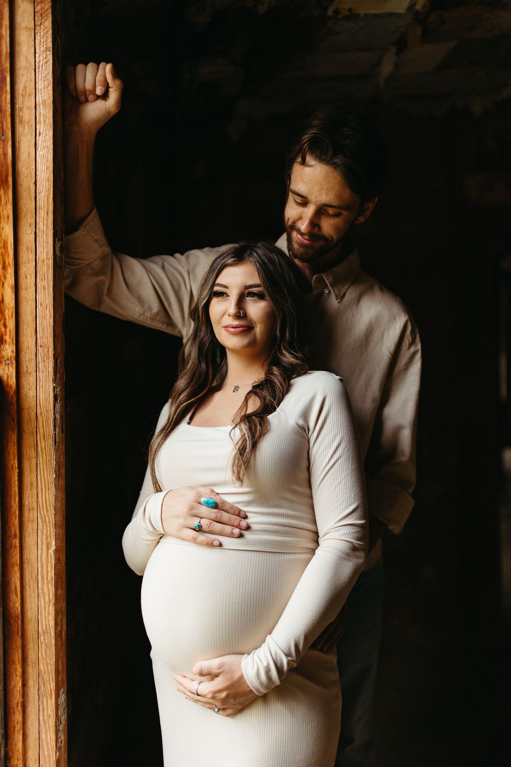Intimate maternity portrait of couple in white clothing standing by wooden doorframe.