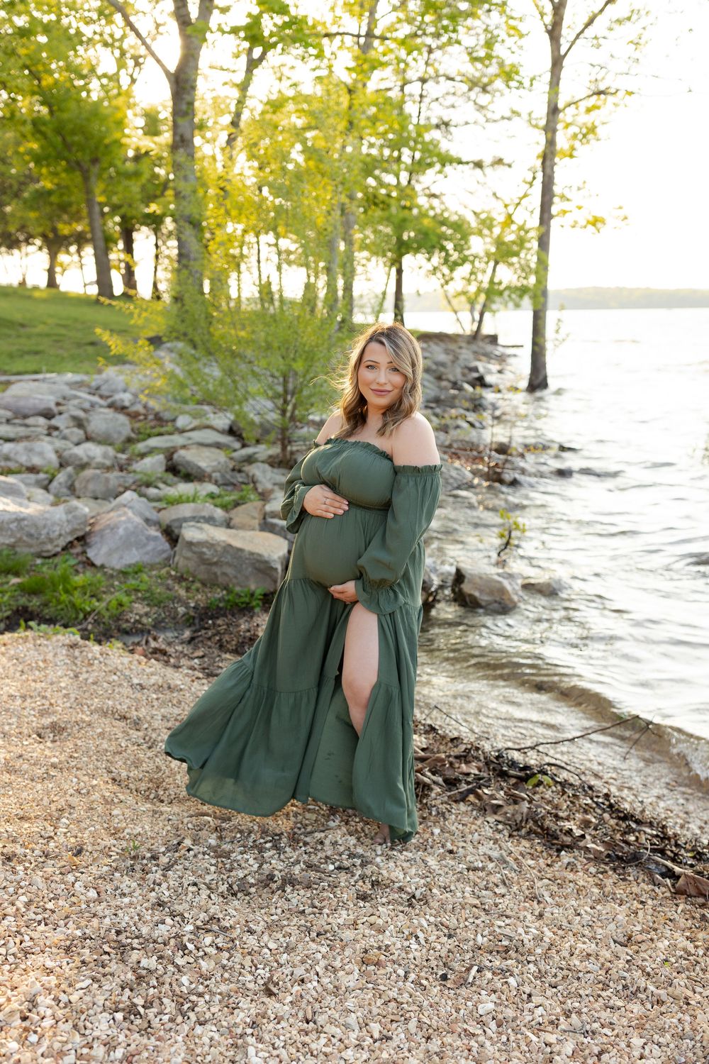 A pregnant woman in a flowing green dress poses by a rocky lakeshore at sunset among trees.