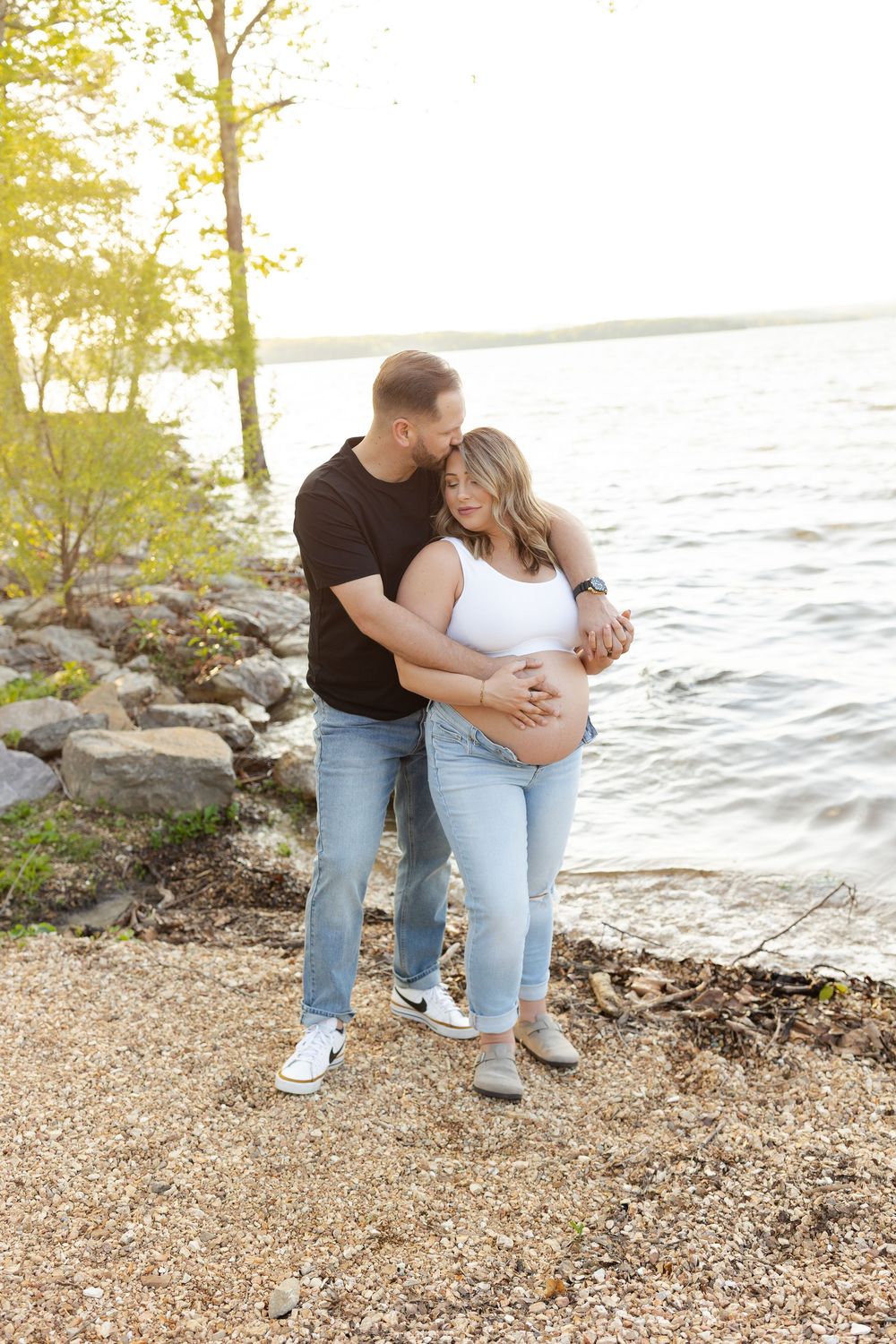 A romantic couple embraces by a lakeside shore during golden hour with trees and rocks in the background.