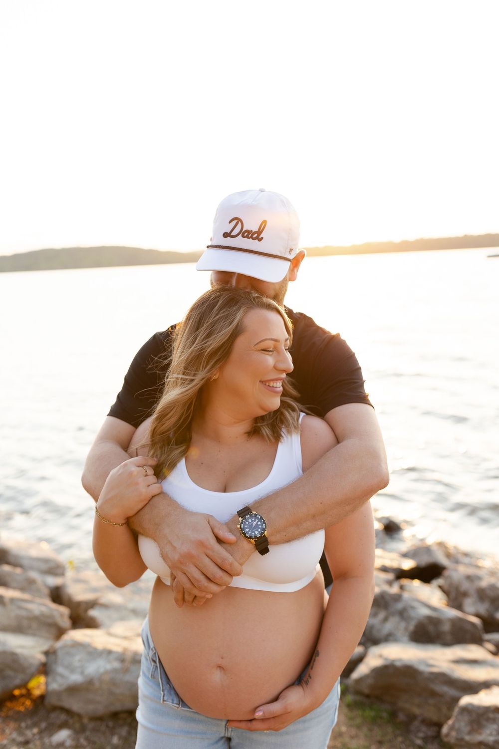 Expecting couple embraces on rocky beach at sunset while wearing casual attire.