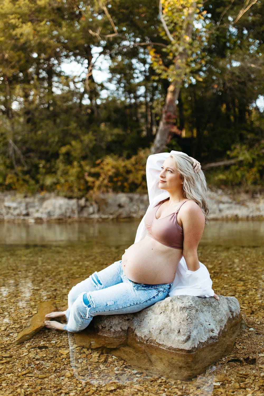 An expectant mother poses on a rock in a river during a sunset photoshoot.