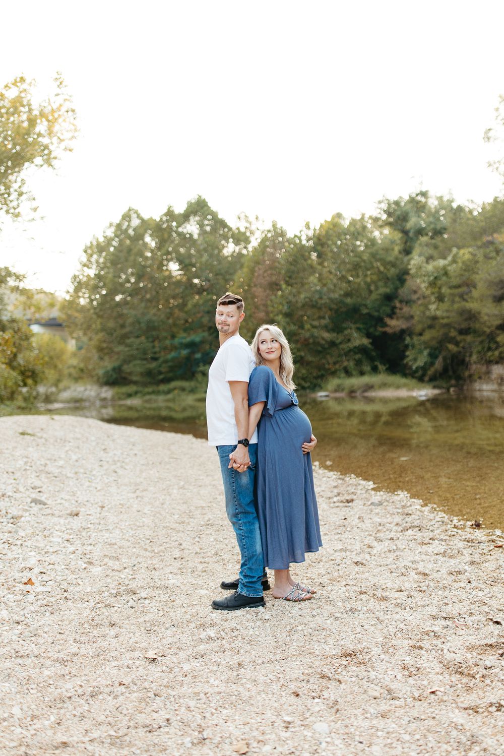 A couple poses back-to-back during their maternity photoshoot by the river.