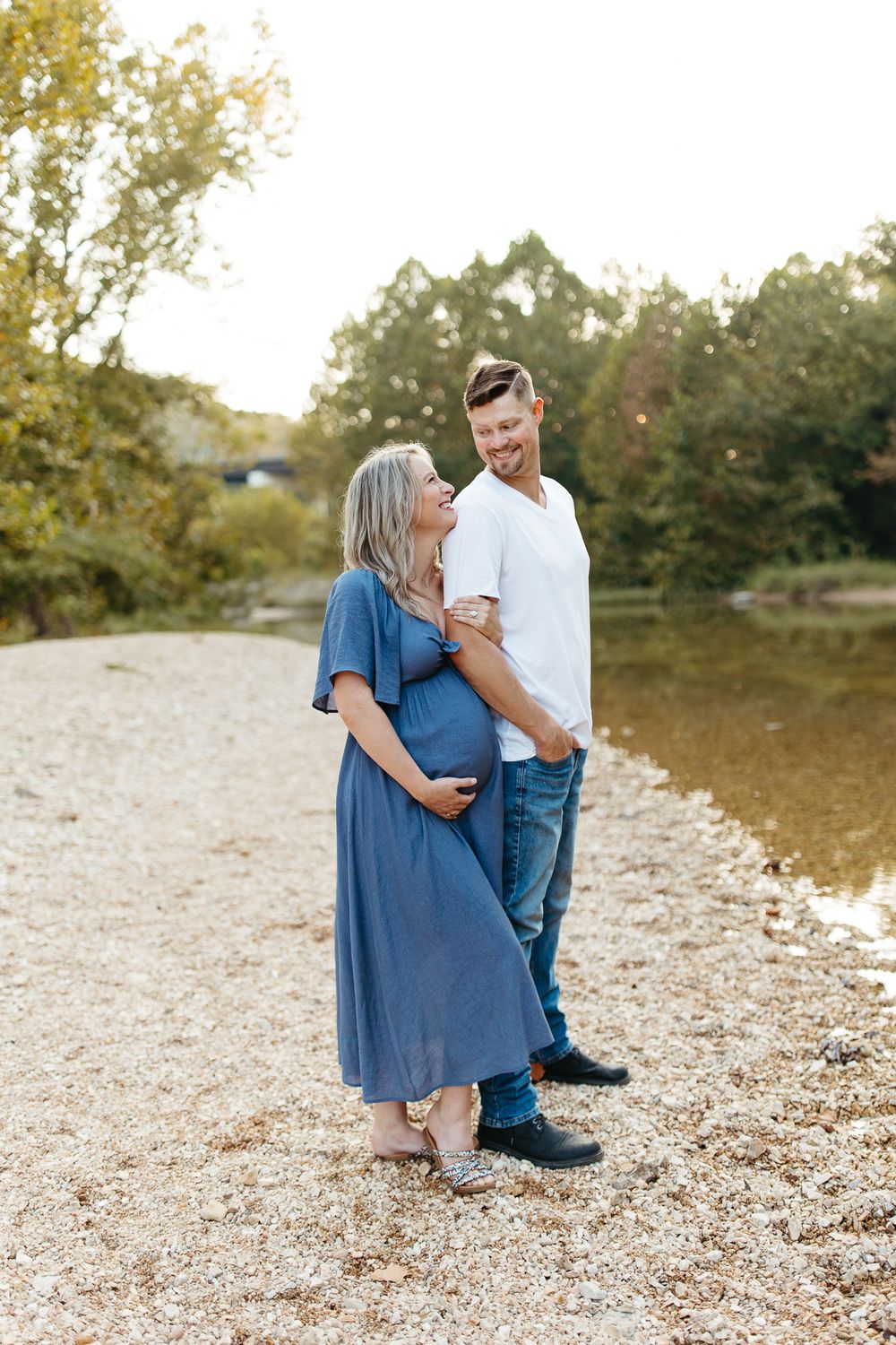 A couple poses together on a riverbank during a sunset maternity session.