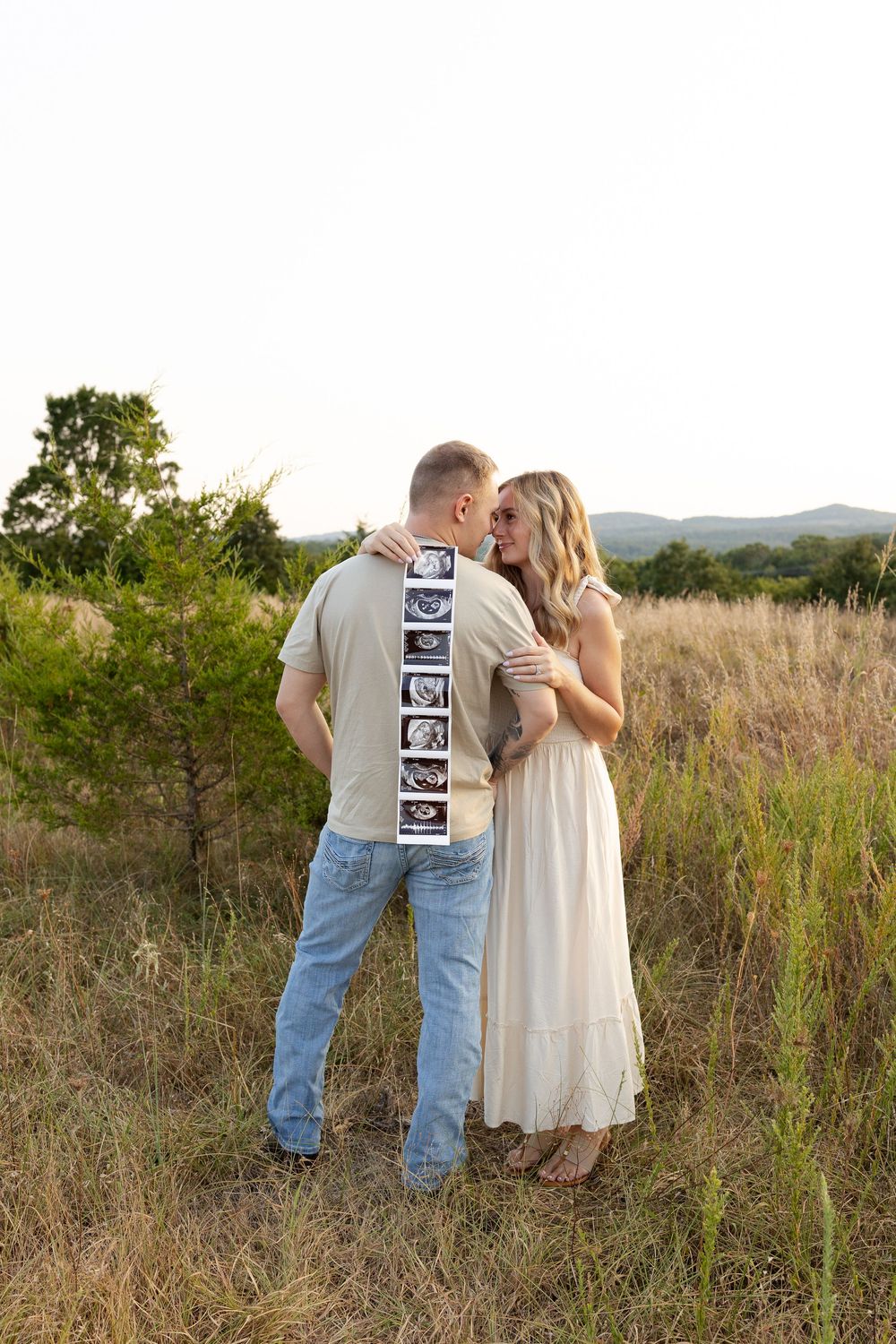 A couple shares a romantic moment in a field while displaying ultrasound photos on the back of a shirt.