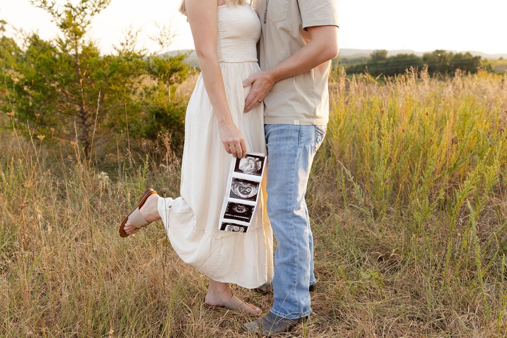 A couple in a field holds ultrasound photos while wearing a white dress and casual attire during a sunset photoshoot.