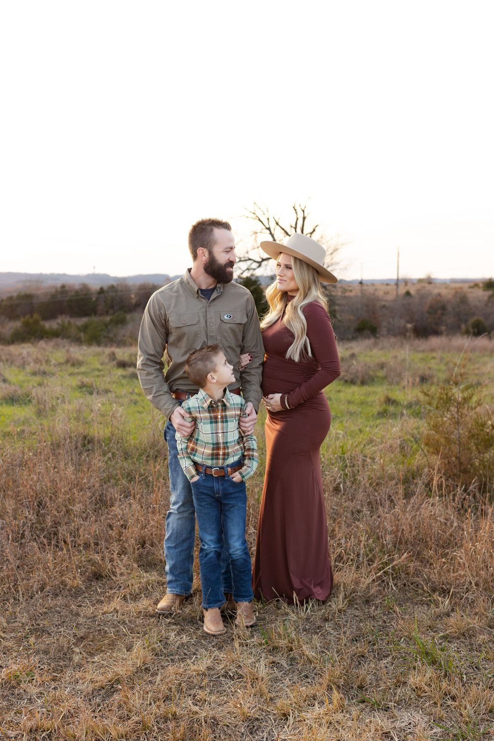 Family maternity photo featuring burgundy dress and plaid shirt in rustic field setting at sunset.