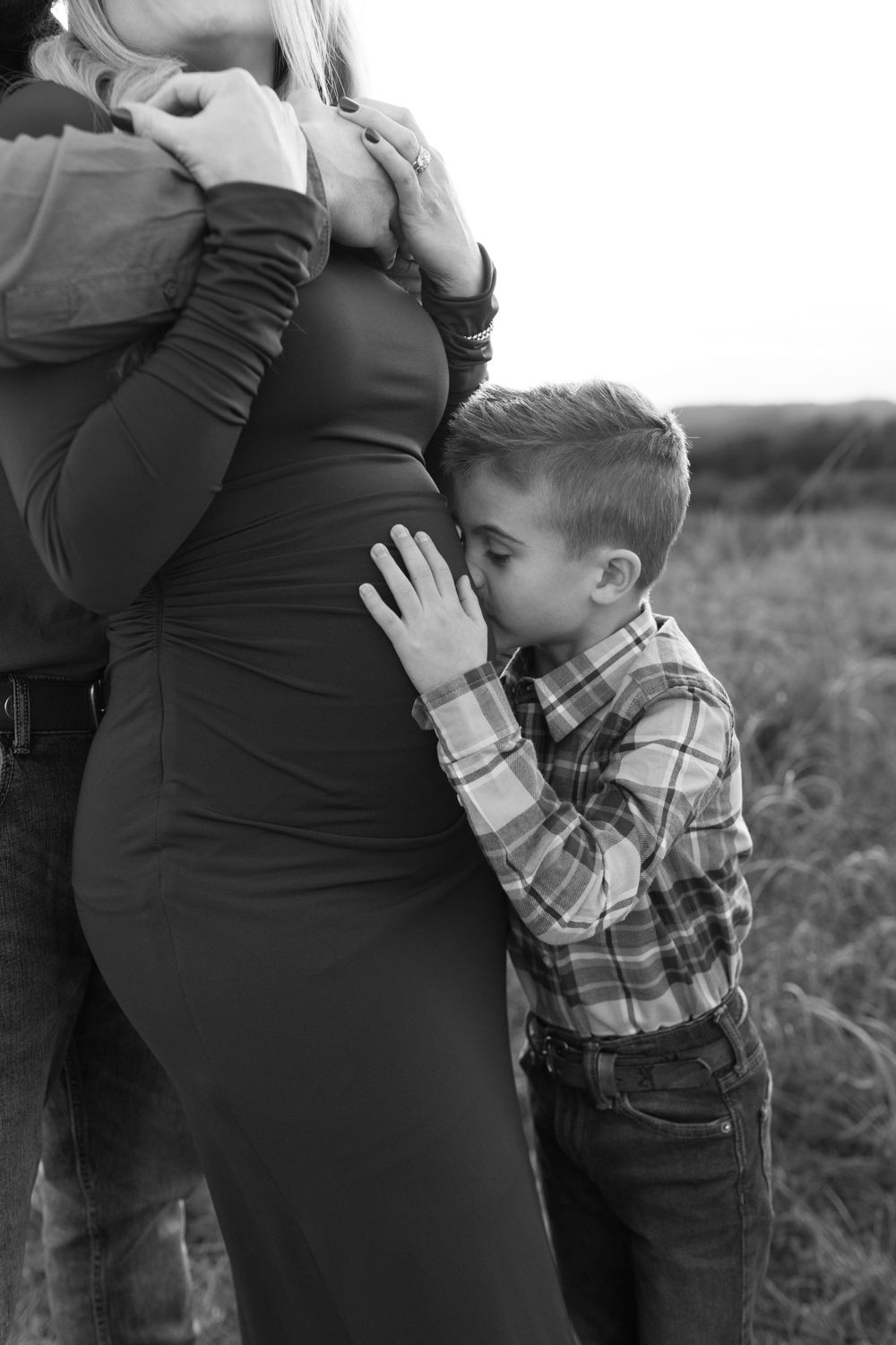 Black and white photo showing tender family moment with pregnant mother in outdoor field.