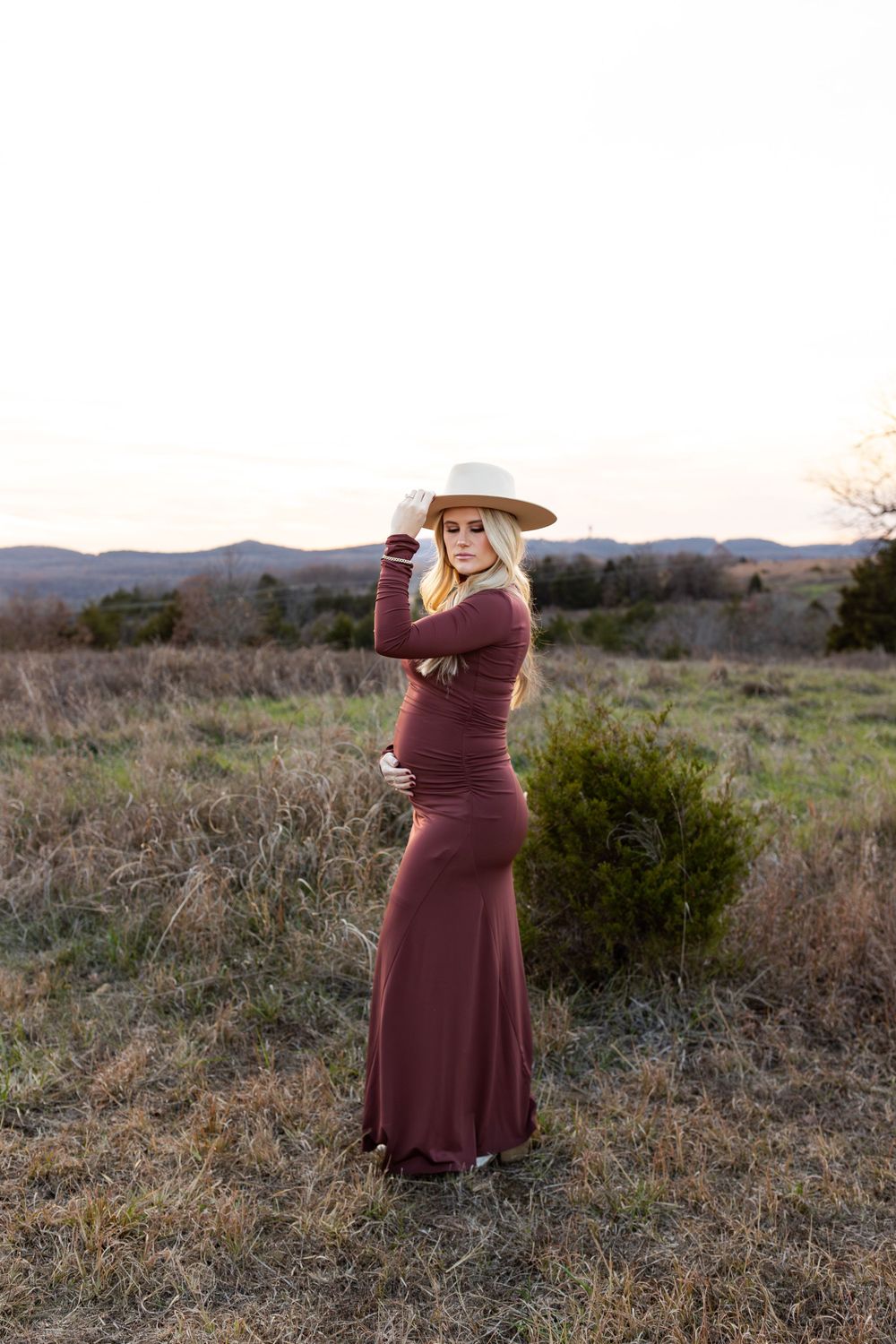 Maternity photo of woman in burgundy dress and wide brim hat posing in field with mountains.