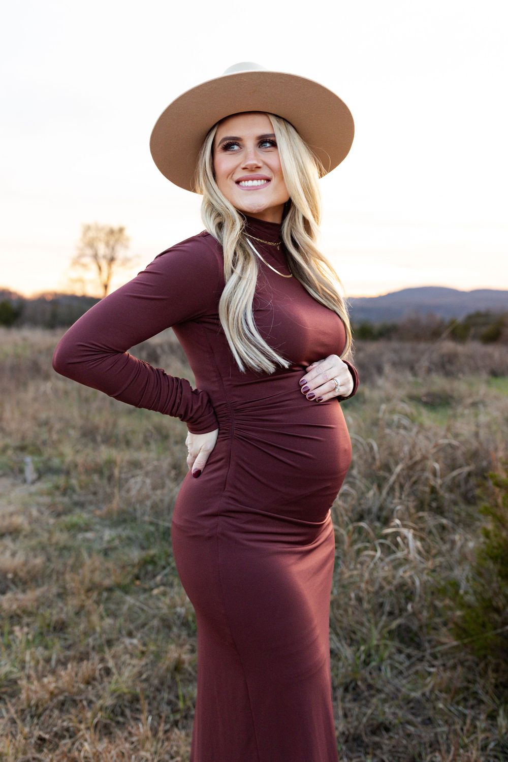 Expectant mother in burgundy dress and tan hat smiling outdoors during golden hour.