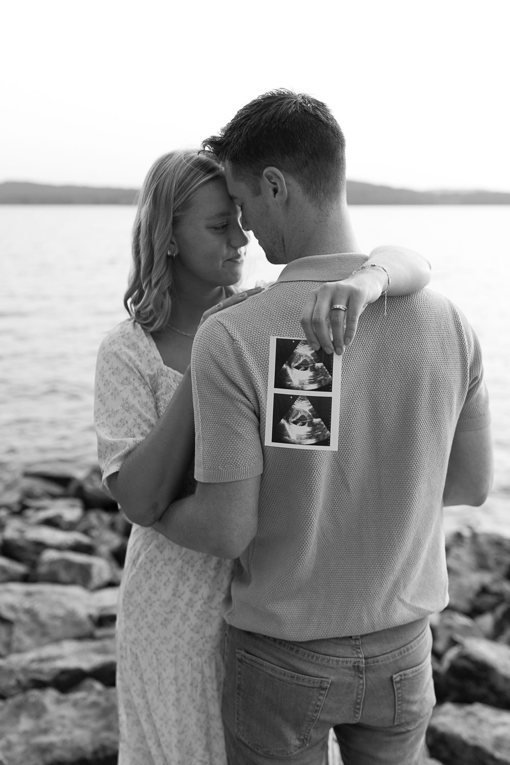 Intimate black and white moment between couple holding ultrasound photo by the water.