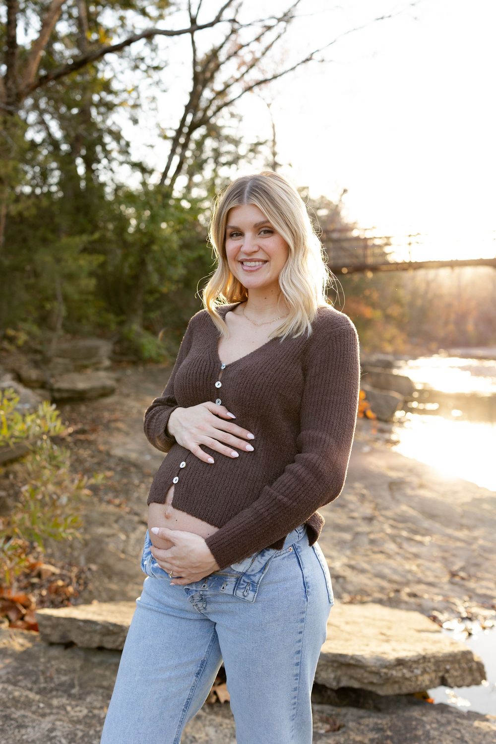 Expectant mother in brown sweater and jeans poses outdoors during golden hour maternity photoshoot.