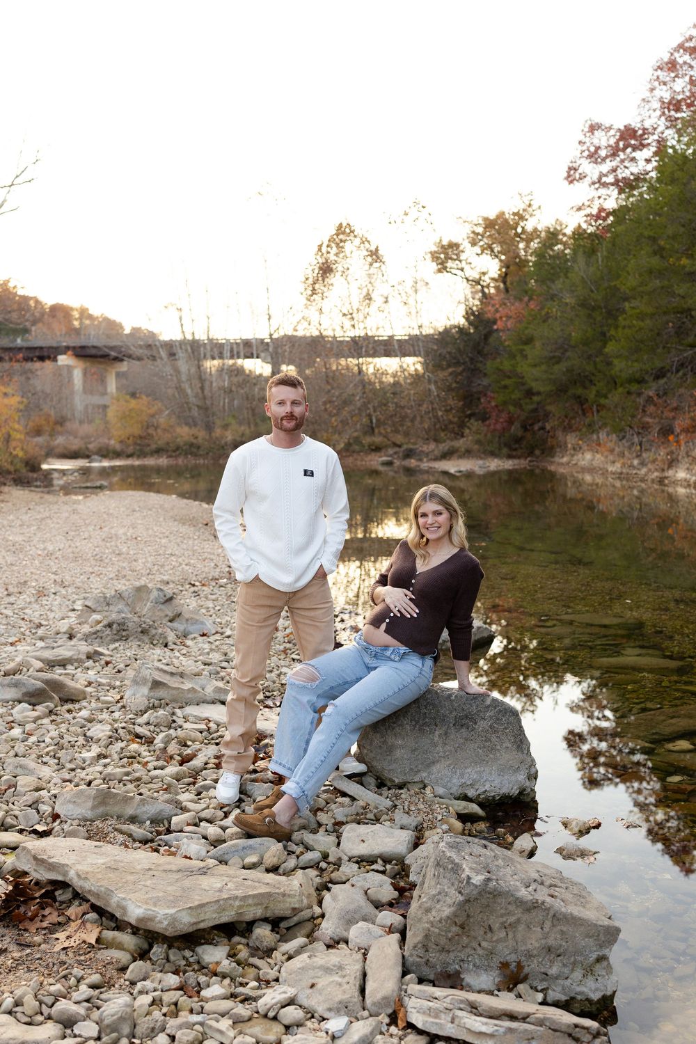 A couple sits together on rocky riverbank during autumn with water and trees in background.