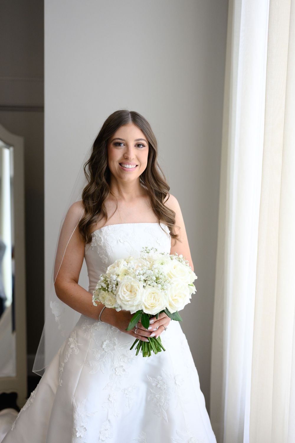 Smiling bride in strapless white lace wedding gown holding elegant white rose bouquet near window.