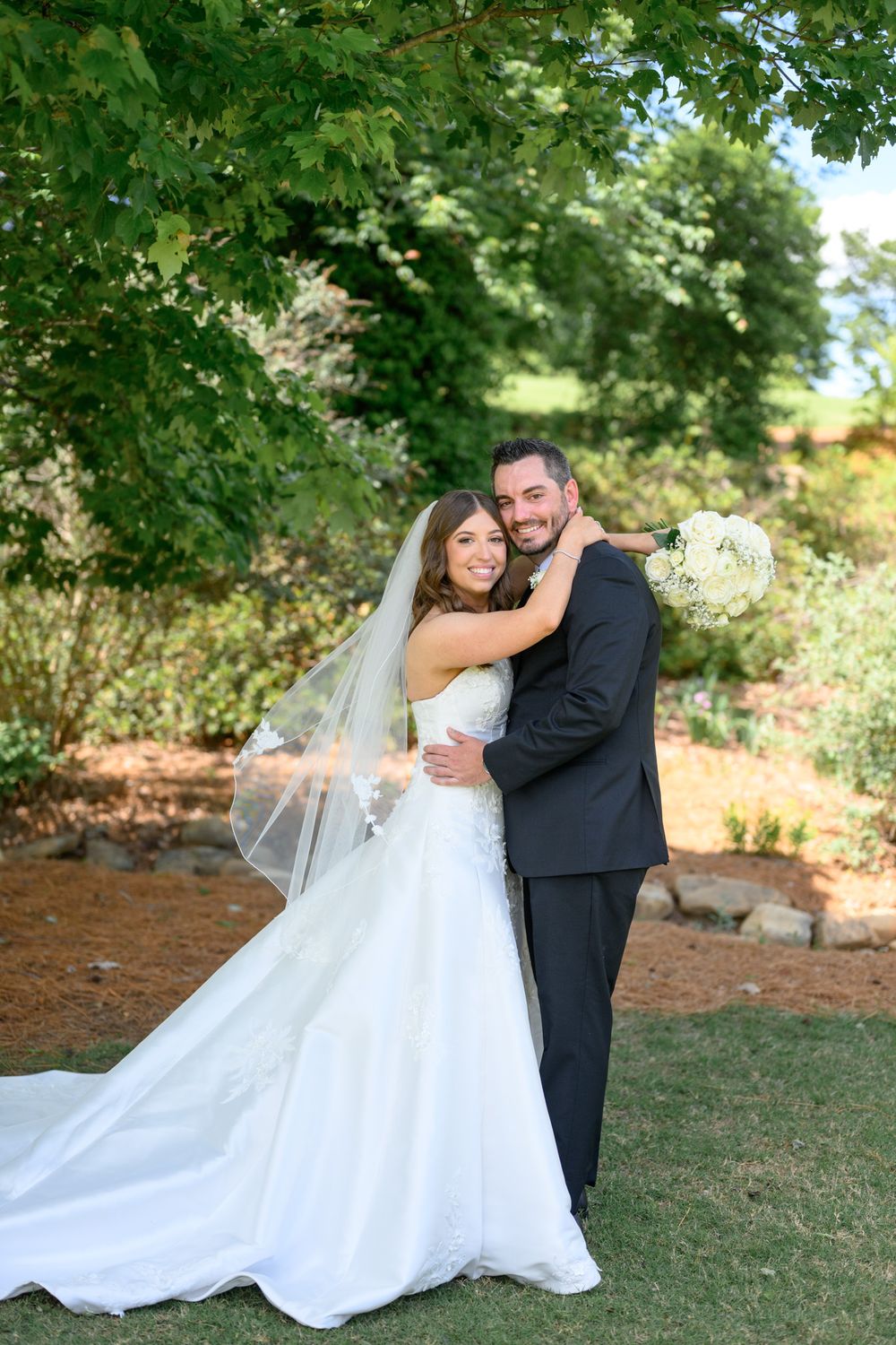 Bride in white gown and veil embraces groom in black suit outdoors in lush garden setting on their wedding day.