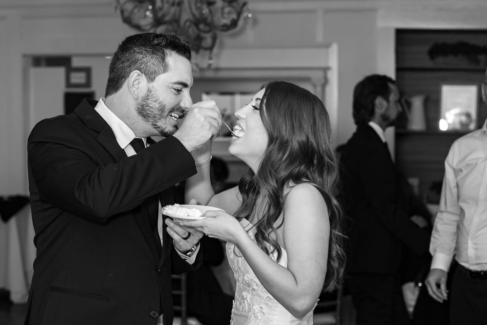 Black and white photo of a groom feeding wedding cake to his bride at their reception.