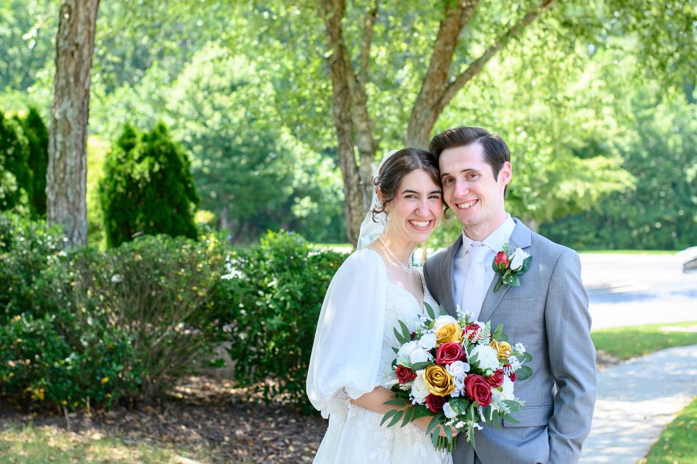Bride and groom smiling together outdoors, holding a colorful bouquet, surrounded by lush green trees on their wedding day.