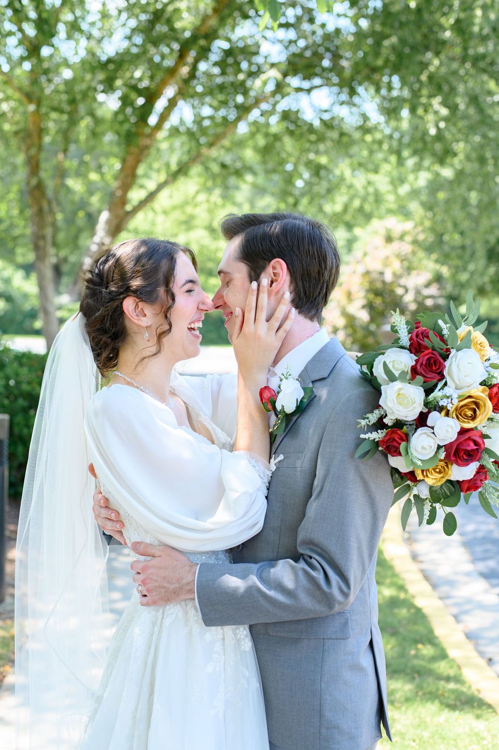 Bride and groom share a joyful moment outdoors, with colorful red and white rose bouquet, surrounded by lush green trees.