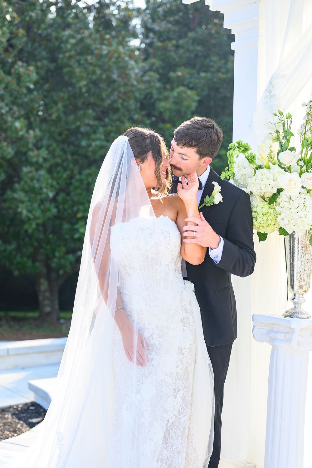 Bride and groom share a romantic kiss at outdoor wedding ceremony with white floral arrangements and columns.