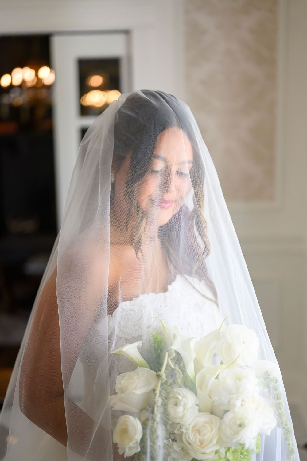 Bride in white lace gown and veil holding elegant white bouquet of roses, looking down in soft indoor light. abbott mansion wedding photos