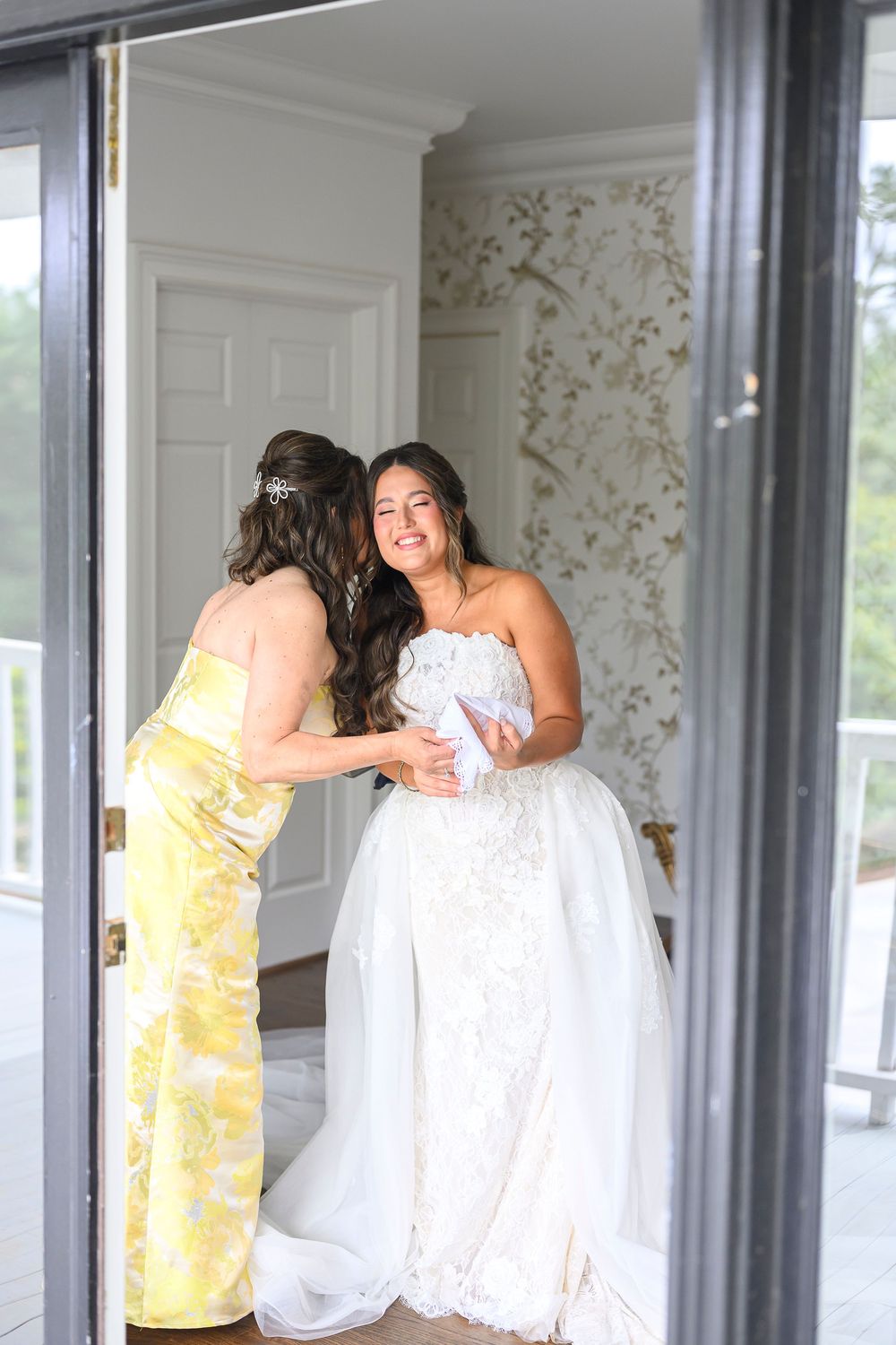 A bride in a white lace gown smiles as her bridesmaid in a yellow floral dress helps her prepare indoors.