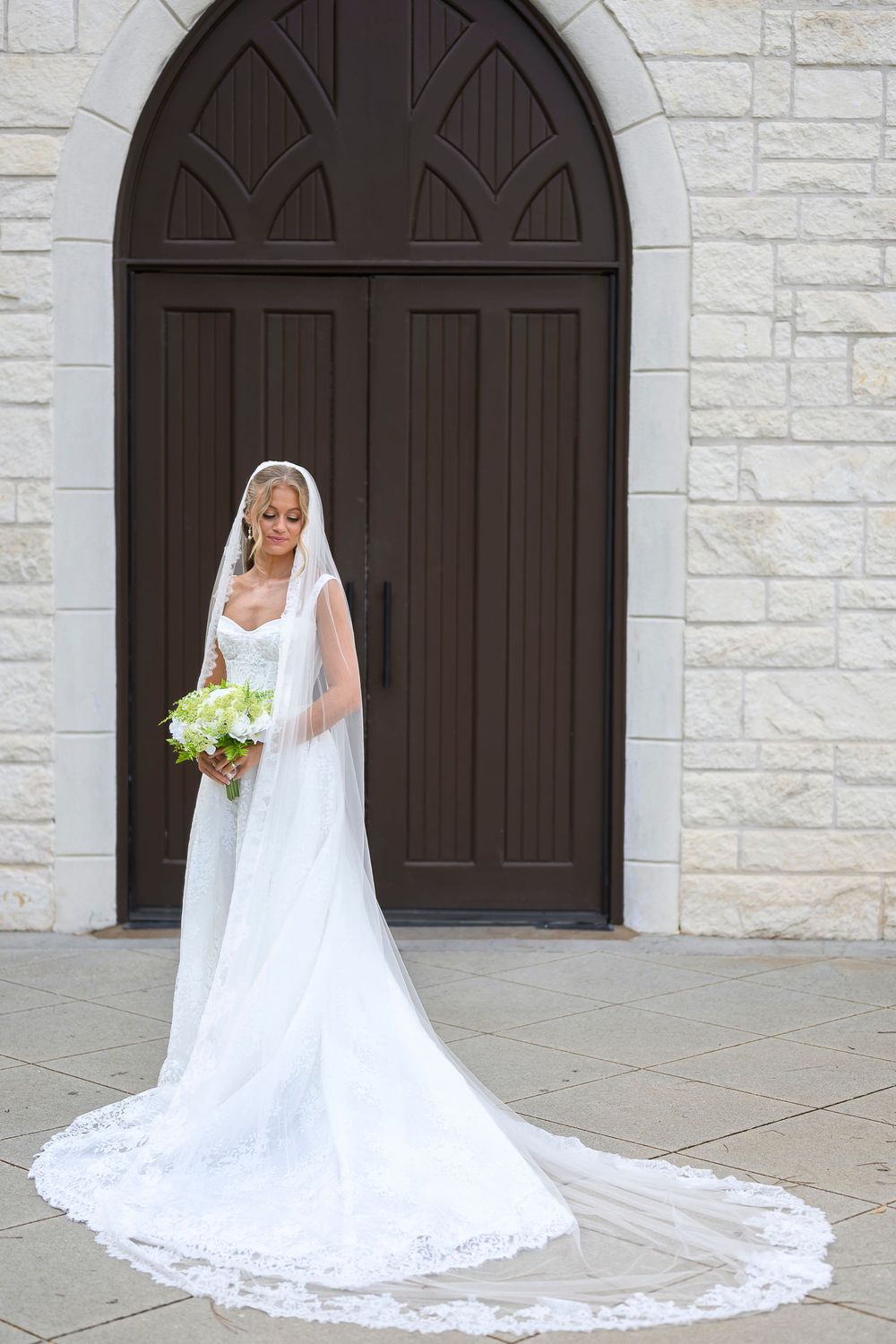 Bride in white gown with long lace veil holding bouquet stands before ornate dark wood church doors.