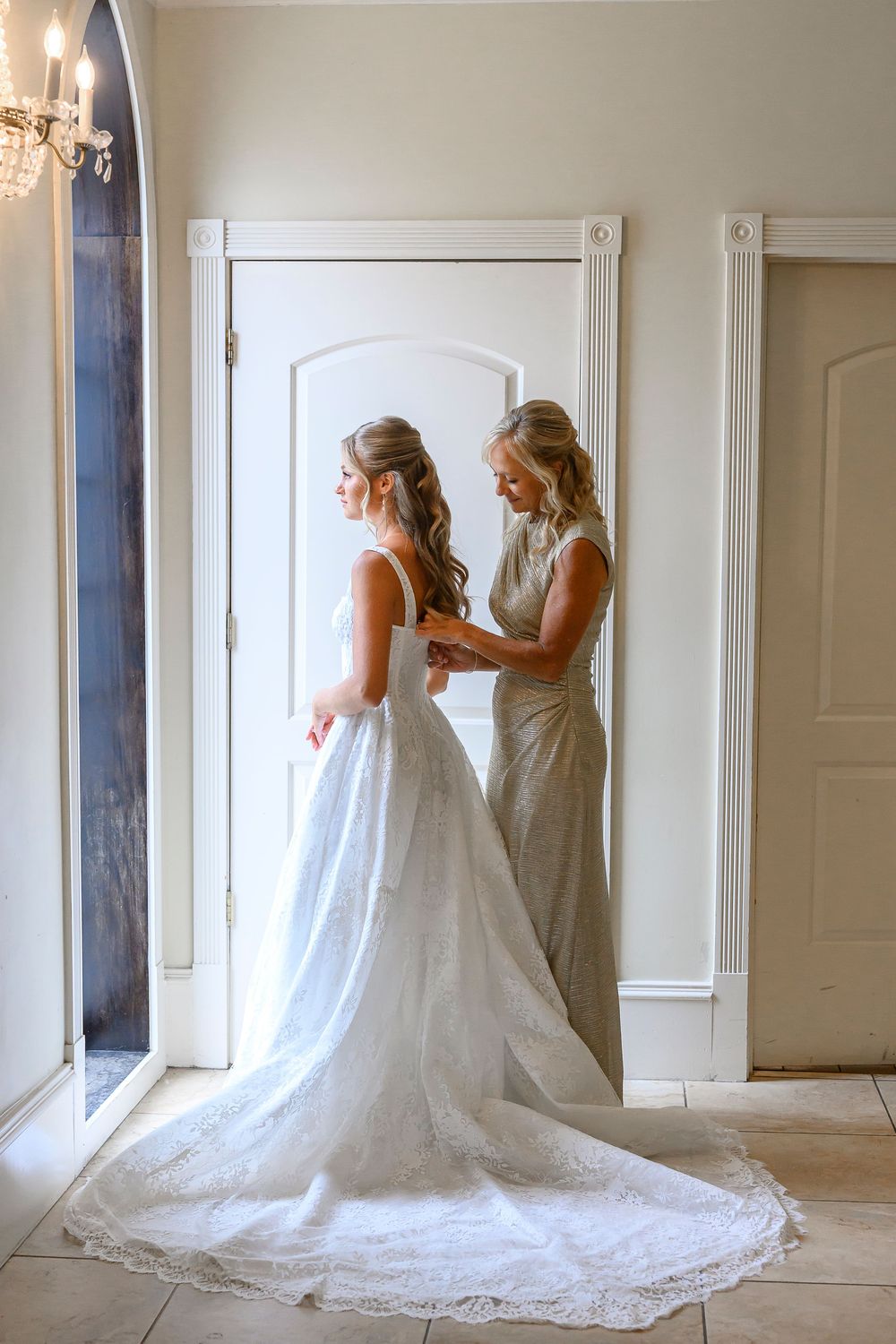 Bride in white lace gown being helped by bridesmaid in silver dress inside elegant venue.