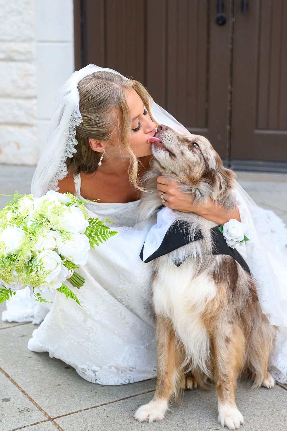 Bride in white lace gown and veil kisses her dog dressed in a tuxedo while holding a white floral bouquet.