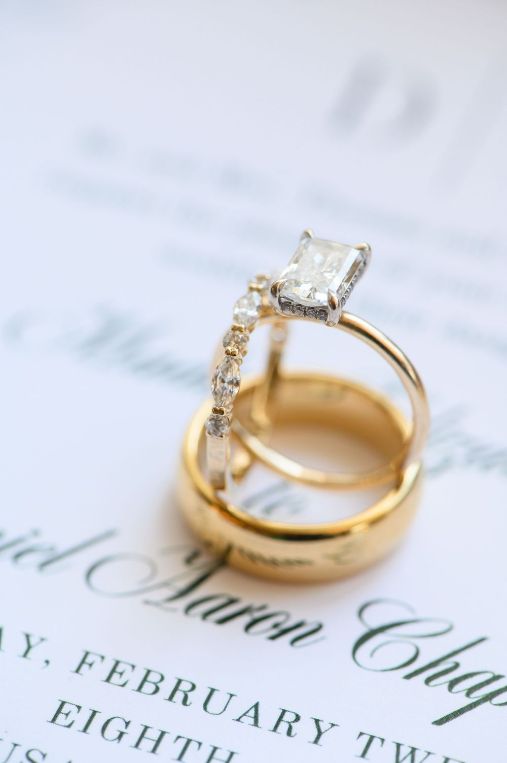 Gold wedding bands and diamond engagement ring stacked on elegant February wedding invitation.