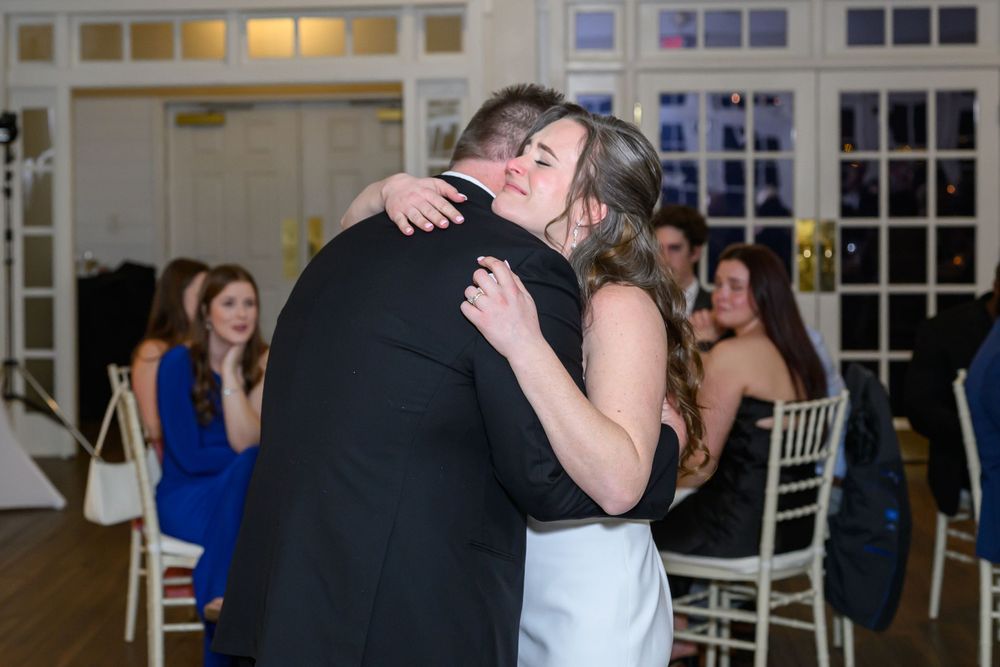 Bride and groom share an emotional first dance hug at elegant wedding reception with guests seated in background.