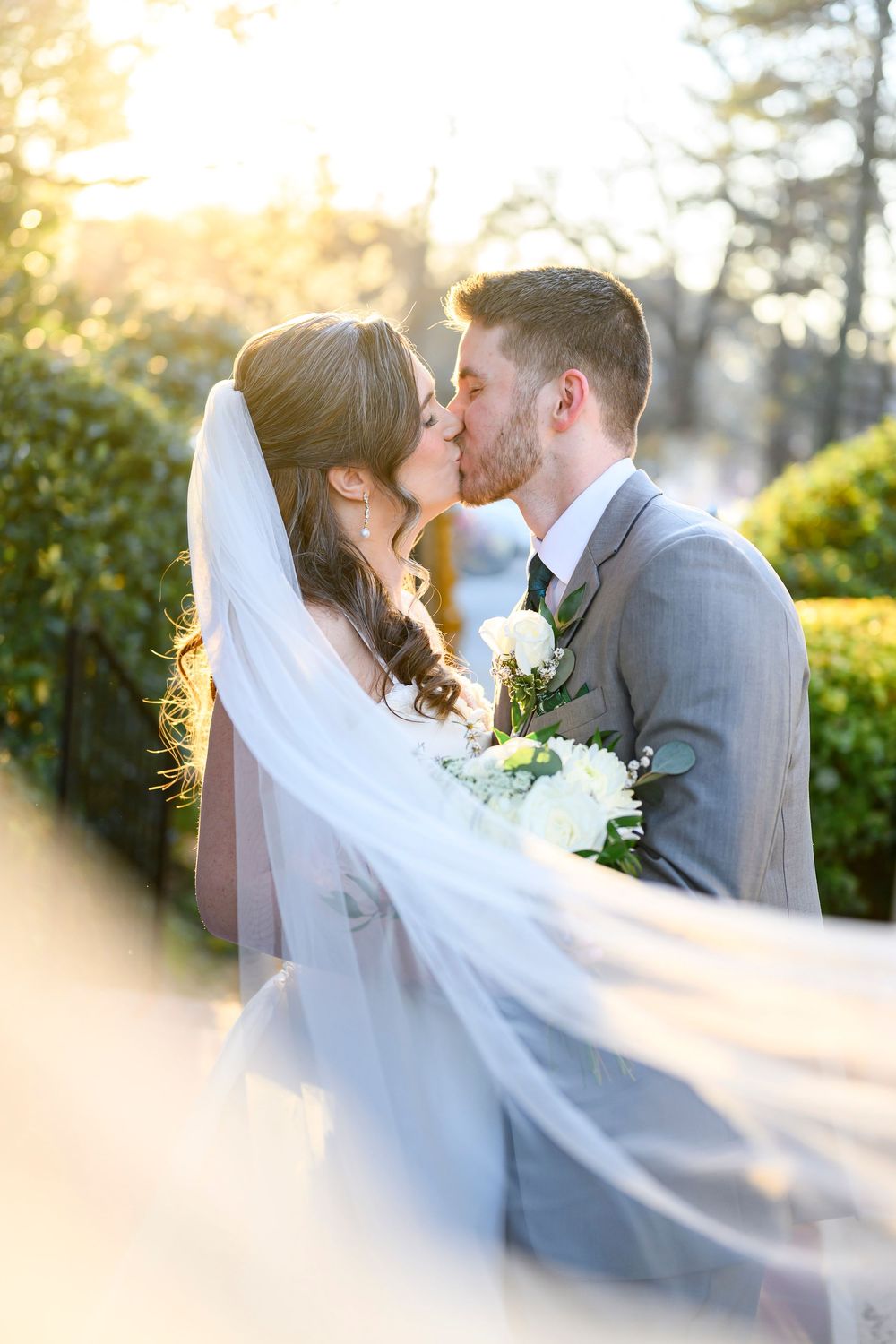 A romantic wedding couple shares a tender kiss in golden sunlight while the bride's veil flows elegantly in the breeze.
