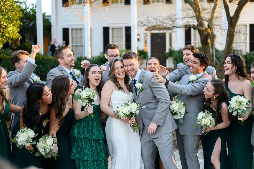 Wedding party wearing emerald green dresses and gray suits laughs together while holding white floral bouquets.