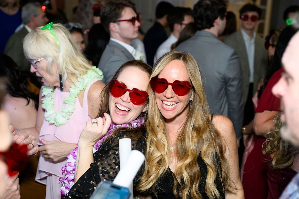 Partygoers wearing red heart-shaped sunglasses smile and celebrate at a festive indoor event.