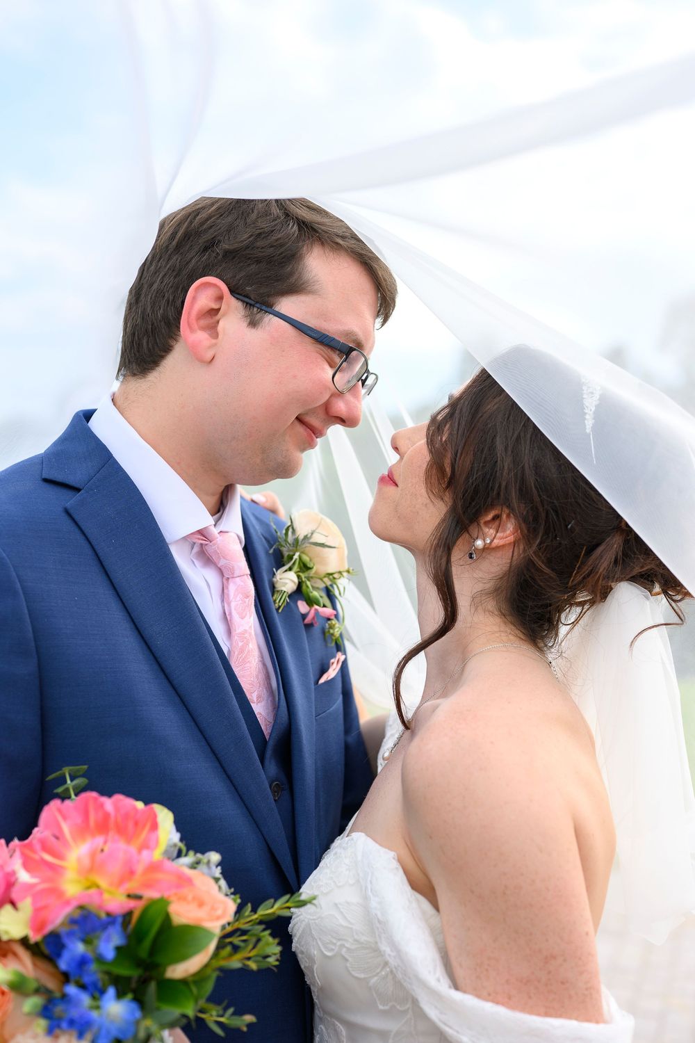 Bride and groom share a romantic moment under a wedding veil, groom in blue suit with pink tie holding colorful bouquet.