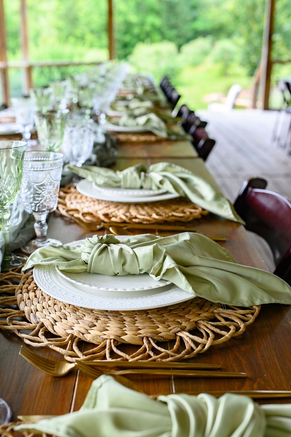 Elegant rustic table setting with woven chargers, white plates, sage green napkins, and crystal glassware for an outdoor event.
