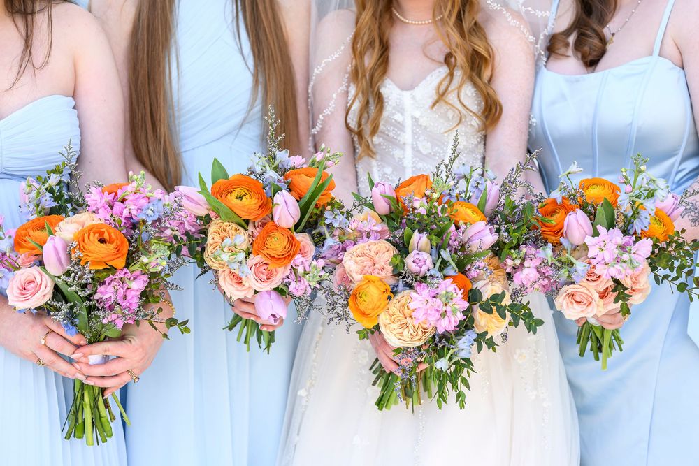 Bride and bridesmaids in light blue dresses holding vibrant orange, pink, and purple wildflower wedding bouquets.