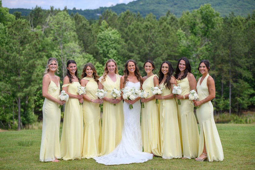 Bride in white gown poses with eight bridesmaids in yellow dresses holding bouquets on a green lawn with trees.