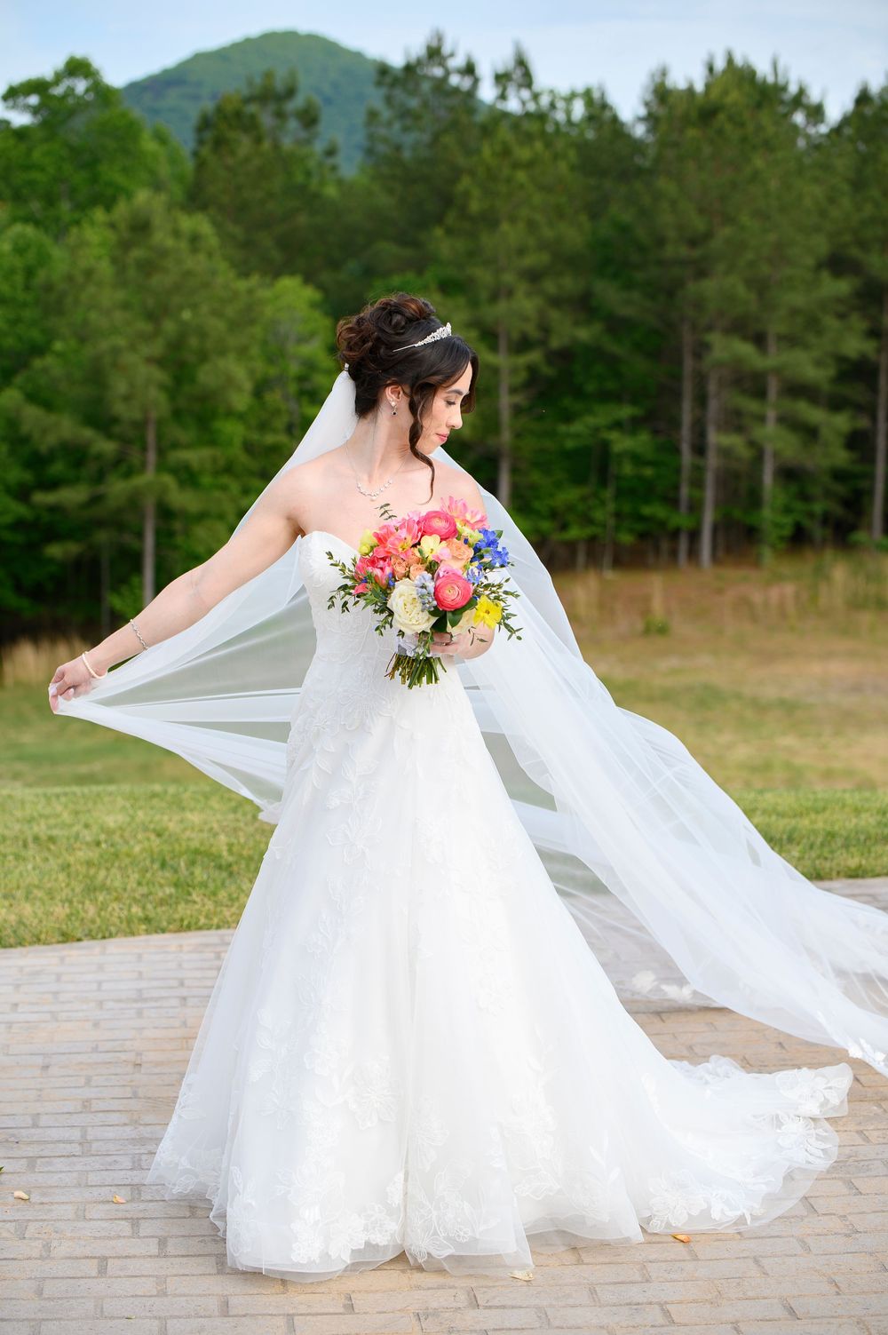 Bride in white lace gown holding colorful bouquet with veil flowing, posing outdoors with green trees and mountain backdrop.
