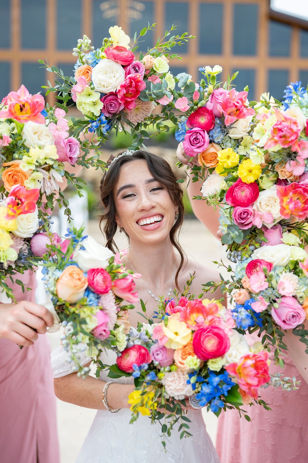 Smiling bride framed by a colorful floral wreath with pink, yellow, and blue flowers on her wedding day.