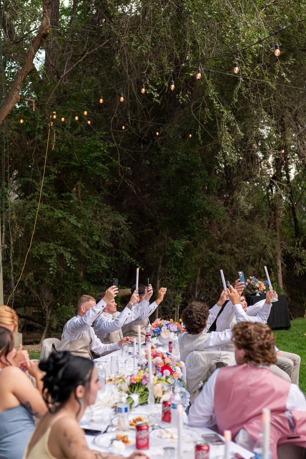 Outdoor dinner party guests raising their hands in celebration under string lights and trees.
