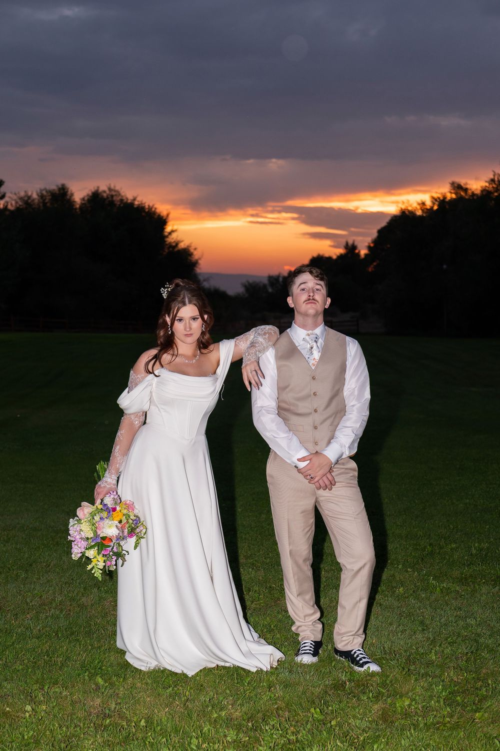 A couple poses playfully in a field at sunset wearing wedding attire and casual sneakers.